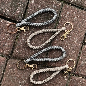 May include: Three handmade wrist keychains with gold-colored hardware. The keychains are made of braided cord in shades of gray and off-white. The keychains are laying on a brick surface.