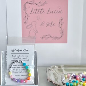 May include: A rainbow-colored beaded bracelet with the name "Lucia" spelled out in white letters. The bracelet is packaged in a white box with a pink card that says "Little Lucia & Me".