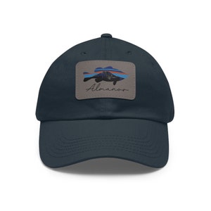 May include: A navy blue baseball cap with a patch featuring a silhouette of a fish with a mountain range inside. The patch is gray with the word "Almanor" written in black cursive.