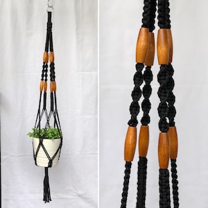 May include: Black macrame plant hanger with wooden beads. The plant hanger is suspended from a metal ring and holds a white pot with a green plant. The design features a tassel at the bottom.