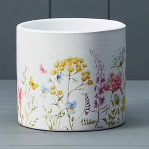 May include: White ceramic plant pot with a vibrant floral design. The design includes yellow, pink, blue, and purple flowers, and butterflies. The cylindrical pot is suitable for indoor or outdoor use. The pot has a matte finish.