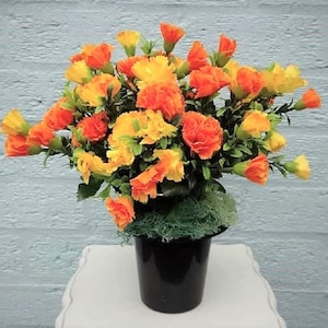 May include: A bouquet of orange and yellow carnations in a black pot. The flowers are arranged in a circular shape and are surrounded by green foliage.