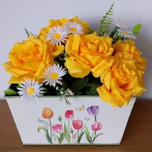May include: A floral display of yellow roses and white daisies in a white rectangular planter. The planter is decorated with illustrations of tulips and butterflies in various colours.