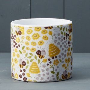 May include: A cylindrical white plant pot decorated with a bee and floral design. The pattern includes yellow sunflowers, brown bees, and stylized beehives on a white backdrop. Suitable for indoor or outdoor use, it measures approximately 15 cm in diameter.