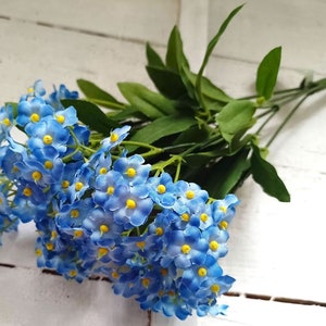 May include: A bunch of artificial blue forget-me-not flowers with yellow centres and green leaves. The flowers are grouped together, creating a full and vibrant display. The stems are long and green, set against a white background.