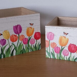May include: Two wooden rectangular boxes with a painted floral design. The boxes are decorated with colourful tulips in shades of pink, orange, yellow, and purple, with green stems and leaves. Butterflies are also painted on the boxes.