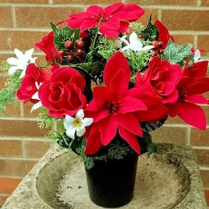 May include: A colourful artificial flower arrangement with red poinsettias, roses, and white blooms. The display is set in a black pot, with green foliage and red berries. The arrangement is placed against a brick wall, creating a festive display.