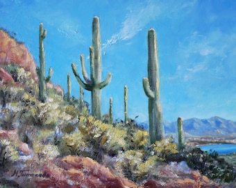 May include: An oil painting depicts a desert landscape with tall saguaro cacti under a bright blue sky. The foreground features reddish-brown rocks and green desert plants. Distant mountains and a lake are visible.