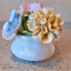 Peut inclure: Un vase en porcelaine blanche avec un bouquet de fleurs. Les fleurs sont jaunes, roses et bleues. Le vase est rond et a une surface lisse.
