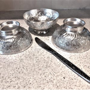 May include: Three silver bowls with an embossed floral design and a black handled knife on a white countertop.