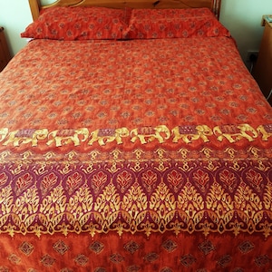 May include: A bed with a wooden headboard and matching orange bedding. The bedding features a repeating pattern of elephants and ornate designs in gold and burgundy. The pillows and comforter are neatly arranged, creating a warm and inviting atmosphere.