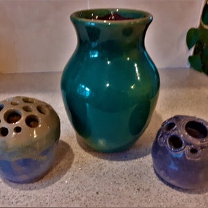 Puede incluir: Tres jarrones de cerámica, uno verde, uno gris azulado y uno marrón y gris azulado. Los jarrones tienen una superficie texturizada y están decorados con agujeros.