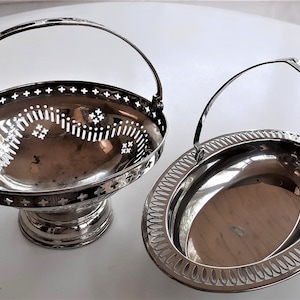 Può includere: Due cesti da portata in argento con manici. Il cesto a sinistra ha un design decorativo traforato e una base a piedistallo. Il cesto a destra ha una base ovale piena con un bordo decorativo.