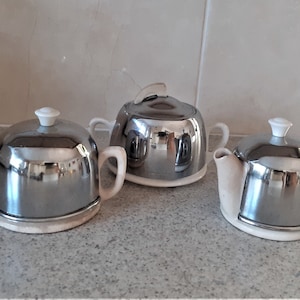 Könnte beinhalten: Ein Vintage-Tee-Set mit Chrom-Finish und weißem Porzellan-Sockel. Das Set beinhaltet eine Teekanne, eine Milchkännchen und eine Zuckerdose.