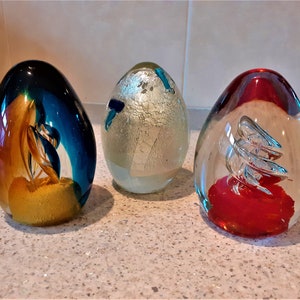 3 glass egg paperweights from the 1980s. 
1  Flamboyant colours in tongues of flame and sapphire
2 Murano glass with silver leaf interior and pieces of blue
3 Silver twist rises from  bright vermillion base 
All 12cm tall, and  under 1kg each.