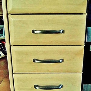 May include: A set of four stacked kitchen drawers featuring a light wood grain finish and beveled edges. Each drawer is equipped with a horizontal, dark metallic handle centered on the front panel, providing a consistent look for the unit.