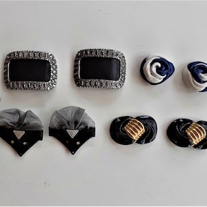 Op de afbeelding: Een assortiment van acht decoratieve haarclips in verschillende stijlen. Twee zilverkleurige clips met zwarte centra, twee zwarte clips met een witte tule strik, twee witte en blauwe stoffen rosetten en twee zwarte clips met een gouden centrum.