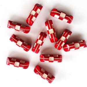 May include: A collection of red and white rectangular beads with a checkerboard pattern. The beads are arranged on a white surface, showcasing their glossy finish and geometric design. These beads could be used for jewelry making or other craft projects.