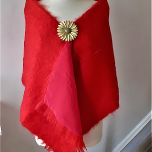 Puede incluir: Un chal de lana roja con borde de flecos y forro rosa. Un broche de flor dorado está prendido al chal.
