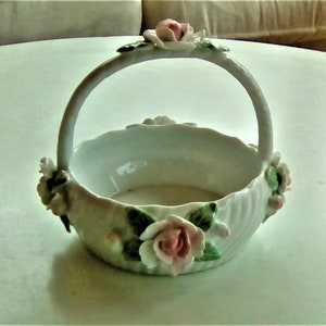 Könnte beinhalten: Ein weißer Porzellan-Korb mit Henkel und floralen Akzenten. Der Korb ist mit rosa und weißen Rosen und grünen Blättern verziert.