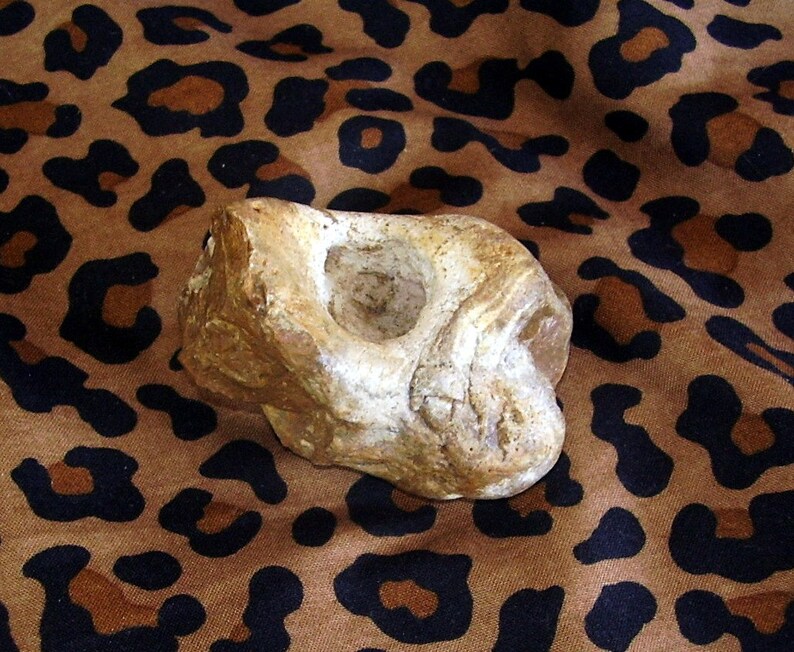 May include: A light brown, irregularly shaped stone with a smooth, round indentation. The stone is resting on a brown and black leopard print fabric.