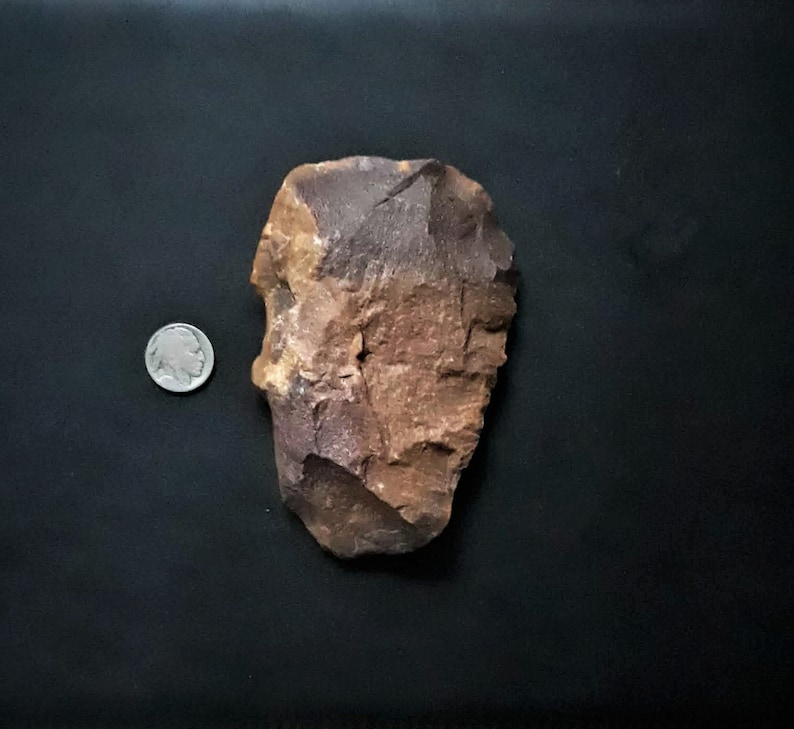 Puede incluir: Una herramienta de piedra grande, marr&oacute;n y roja con una superficie rugosa y astillada. La herramienta es aproximadamente del tama&ntilde;o de una moneda de plata.