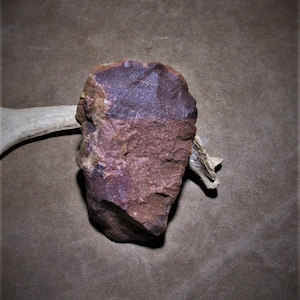 Puede incluir: Una herramienta de piedra de color marr&oacute;n rojizo con una forma irregular y rugosa. La piedra tiene un aspecto moteado con &aacute;reas de color p&uacute;rpura y marr&oacute;n oscuro.