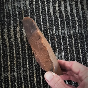 Puede incluir: Una herramienta de piedra marr&oacute;n, lisa y ligeramente curvada. La piedra tiene un extremo puntiagudo y una base redondeada. Es probable que sea una herramienta de mano utilizada para raspar o cortar.
