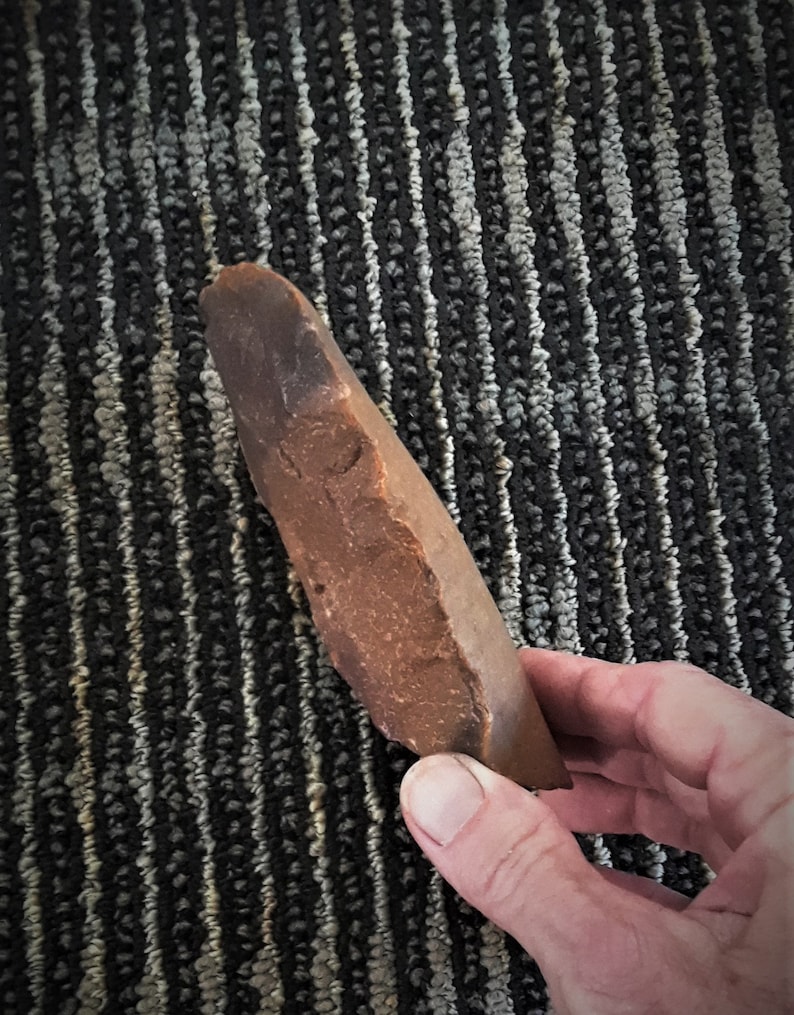 Puede incluir: Una herramienta de piedra marr&oacute;n, lisa y ligeramente curvada. La piedra tiene un extremo puntiagudo y una base redondeada. Es probable que sea una herramienta de mano utilizada para raspar o cortar.