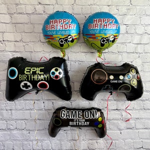 Op de afbeelding: Vier zwarte en blauwe ballonnen met een gamethema. Twee ballonnen zijn rond en zeggen "Happy Birthday You've Leveled Up!" Twee ballonnen zijn gevormd als gamecontrollers. Eén zegt "Epic Birthday!" en de andere zegt "Game On! It's Your Birthday!"