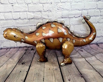 Pode incluir: Um balão de dinossauro inflável marrom e dourado em forma de Anquilossauro. O balão tem um dorso espinhoso e uma cauda longa.