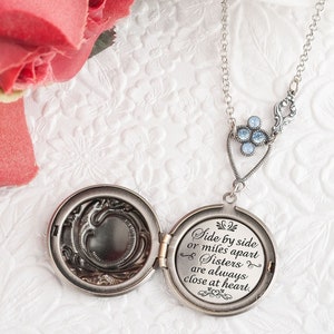 May include: Silver locket necklace with a blue crystal charm. The locket opens to reveal a blank space for a photo. The back of the locket has an inscription that reads "Side by side or miles apart Sisters are always close at heart."