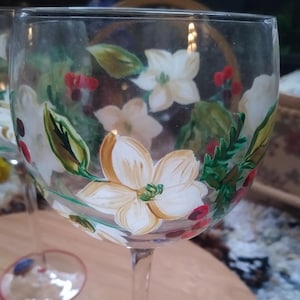 May include: Clear wine glass with a hand-painted floral design. The glass features white magnolia flowers, red berries, and green leaves. The base of the glass has a red rim. The stem is clear.