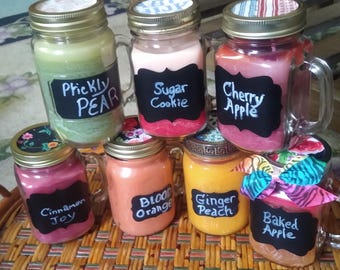 May include: Seven glass mason jar candles with handles and metal lids. Each jar features a black chalkboard-style label with handwritten text: Prickly Pear, Sugar Cookie, Cherry Apple, Cinnamon Joy, Blood Orange, Ginger Peach, and Baked Apple.