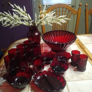 May include: A selection of ruby red glassware, featuring a large bowl, smaller bowls, drinking glasses, and a divided serving dish. A vase with white floral stems is also visible, forming a vintage dining set.