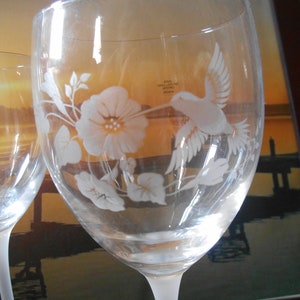 May include: Two clear glass wine glasses with etched designs of a hummingbird and flowers. The glasses have a frosted base and are etched with the text "#2000 GALLI-LUXE INC. VENICE, FL"