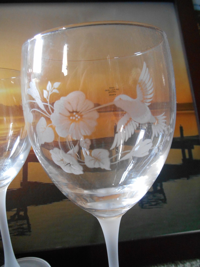 May include: Two clear glass wine glasses with etched designs of a hummingbird and flowers. The glasses have a frosted base and are etched with the text "#2000 GALLI-LUXE INC. VENICE, FL"