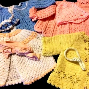 May include: A collection of handmade crochet baby clothes in various pastel colors. The assortment includes sweaters, hats, and headbands in shades of blue, pink, yellow, and white. The garments feature intricate stitch patterns and decorative edging.