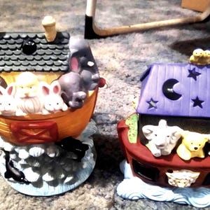 May include: Two ceramic Noah's Ark figurines. One ark is orange with a giraffe, lion, rabbits, and an elephant. The other ark is red with a purple roof, a moon, stars, and animals. The arks sit on a base of white waves.