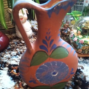 May include: A terracotta-colored pitcher with a handle and spout. The pitcher is decorated with blue floral designs and green leaves. The rim and handle are accented with blue paint. The pitcher is sitting on a patterned rug.