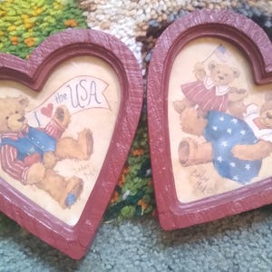Puede incluir: Dos marcos en forma de coraz&oacute;n con ilustraciones de osos de peluche. Un marco presenta un oso con una pancarta que dice "I love the USA". El otro marco muestra dos osos, uno de ellos sosteniendo una bandera. Los marcos est&aacute;n pintados de rojo.