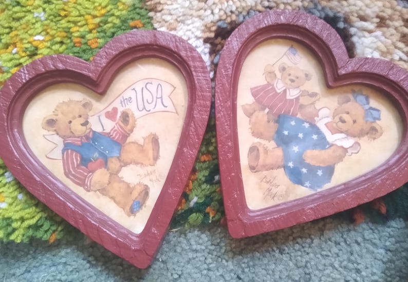 Puede incluir: Dos marcos en forma de coraz&oacute;n con ilustraciones de osos de peluche. Un marco presenta un oso con una pancarta que dice "I love the USA". El otro marco muestra dos osos, uno de ellos sosteniendo una bandera. Los marcos est&aacute;n pintados de rojo.