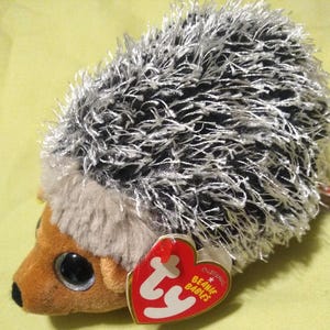 May include: A plush hedgehog toy with a brown face, large black eyes, and a red heart-shaped tag. The hedgehog's back is covered in black and white spiky fur. The tag reads "Original Beanie Babies".