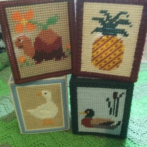 May include: Four square tissue box covers with different embroidered designs. The first cover features a brown turtle with a green leaf and a pink flower. The second cover features a yellow pineapple with green leaves. The third cover features a white duck. The fourth cover features a brown duck with green reeds.