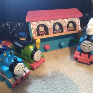 May include: A wooden train station playset with three train engine figures inside the windows. There are three additional train engines on the floor, one blue, one green, and one red.