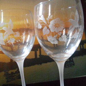 May include: Two clear glass wine glasses with frosted stems and etched designs of hummingbirds and flowers. The glasses are on a dark surface.