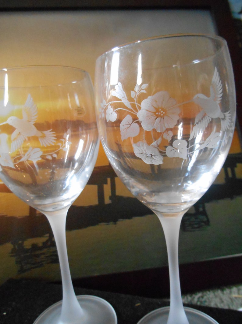 May include: Two clear glass wine glasses with frosted stems and etched designs of hummingbirds and flowers. The glasses are on a dark surface.