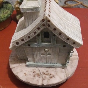 Puede incluir: Figurita decorativa en miniatura con forma de casa, de color azul claro y blanco. La casa tiene una chimenea, una peque&ntilde;a ventana y una puerta. El tejado est&aacute; hecho de tablones de madera y adornado con peque&ntilde;os clavos marrones. La figurita est&aacute; sobre una base redonda y texturizada.