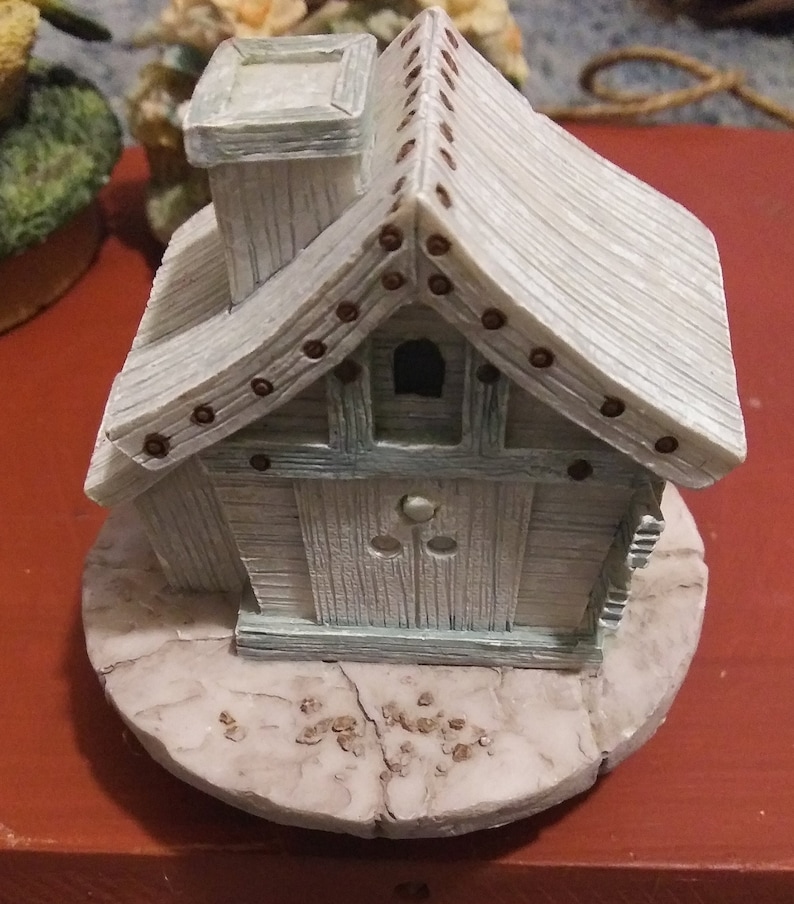Puede incluir: Figurita decorativa en miniatura con forma de casa, de color azul claro y blanco. La casa tiene una chimenea, una peque&ntilde;a ventana y una puerta. El tejado est&aacute; hecho de tablones de madera y adornado con peque&ntilde;os clavos marrones. La figurita est&aacute; sobre una base redonda y texturizada.