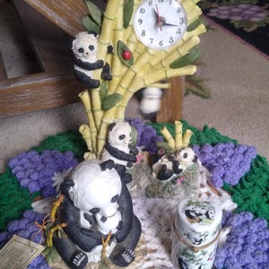May include: A set of four panda figurines, including a clock with a panda climbing a bamboo stalk, a panda family, and a panda holding a baby panda. The figurines are on a white and purple crocheted doily.